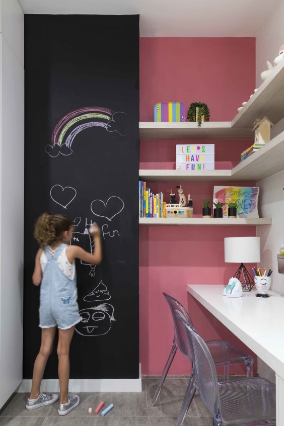 Girl’s bedroom with pink wall paint and black blackboard for drawing on next to to a white desk with with shelves